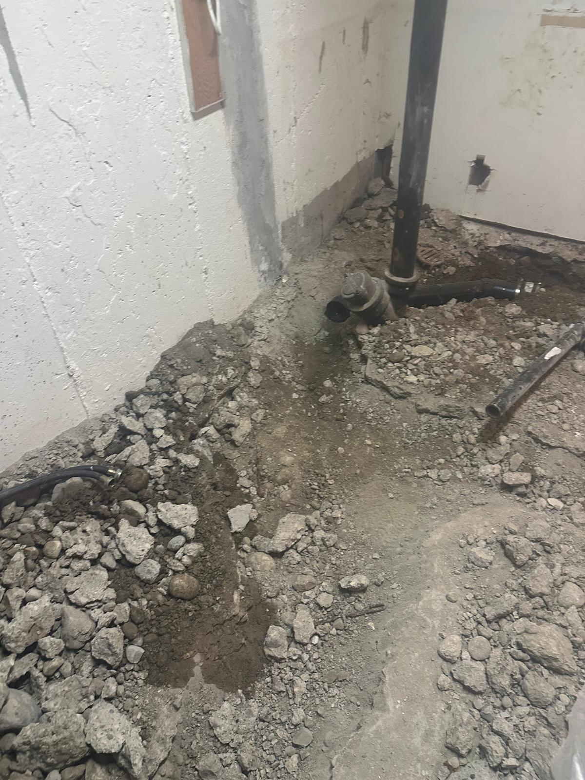 Basement slab demolition with concrete chunks broken out around cast iron stack and plumbing rough-in for new bathroom drain lines