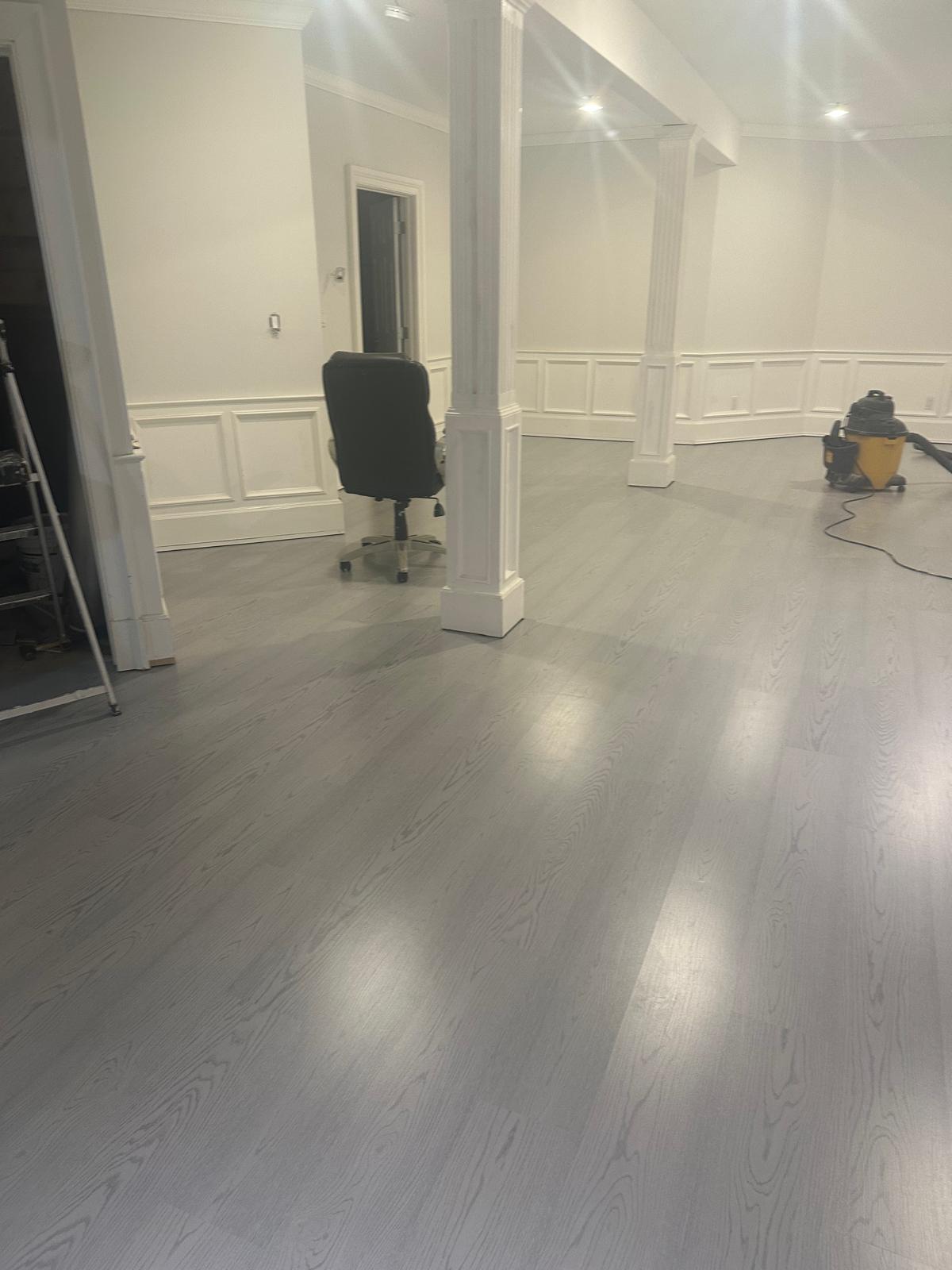 Grey luxury vinyl plank flooring installed throughout an open finished basement with white wainscoting and columns